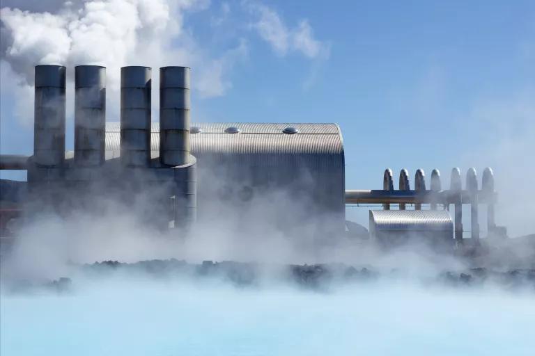 Steam rises off blue water in front of a power plant with four stacks