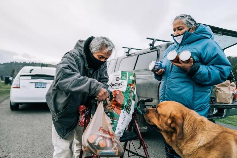 A man wearing a face mask leans on a walker and holds a plastic bag full of groceries while a woman in a face mask holds canned food, and a dog looks on