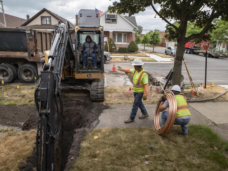A construction worker sits in an excavator that’s digging a hole in a front yard of a home to replace its lead water service line, while one worker stands nearby and another worker is crouched on the side, holding new pipes that would replace the lead ones.