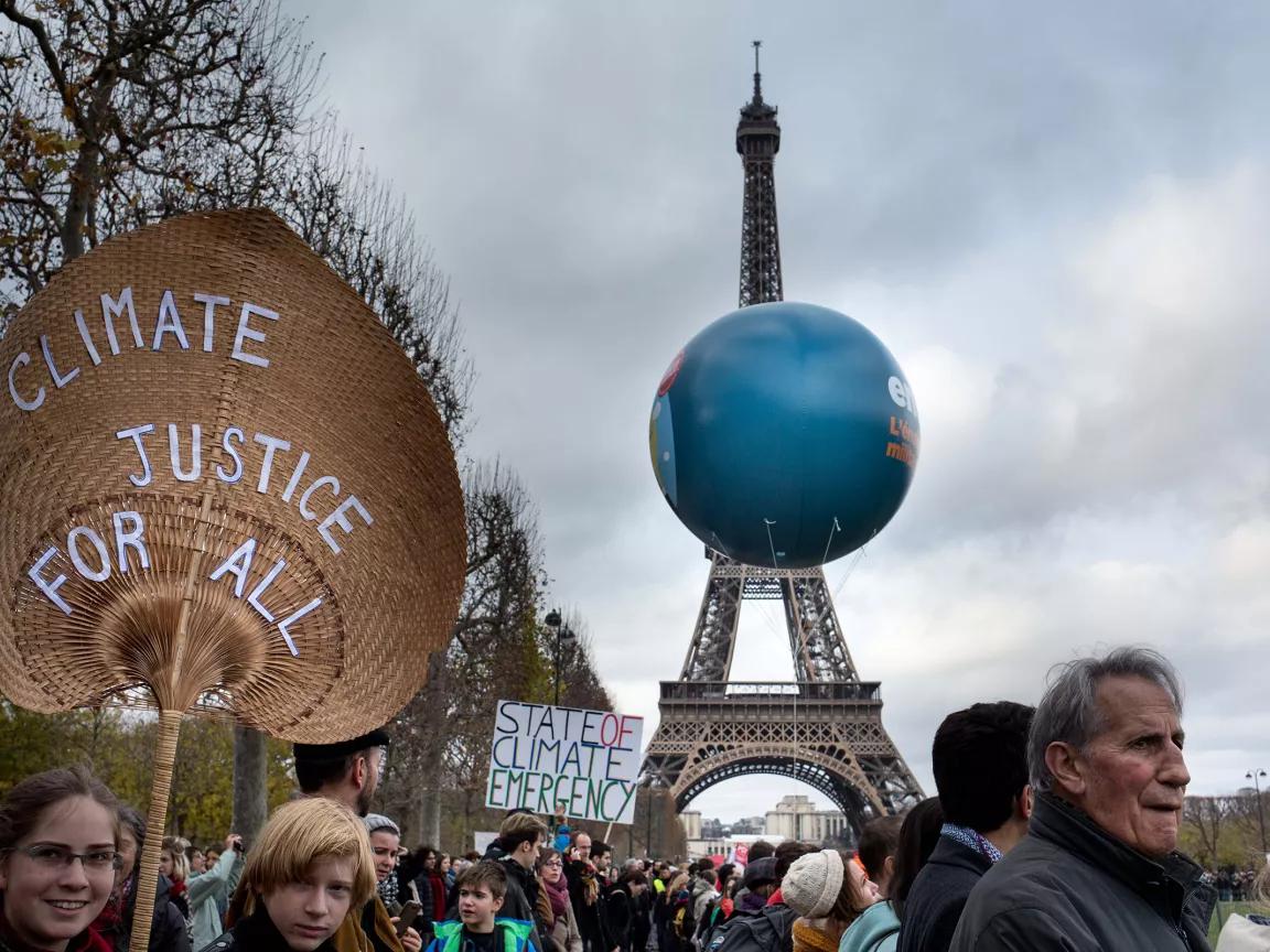 With the Eiffel Tower in the background, a crowd of people protests. One person holds a sign with the words "Climate Justice for All".