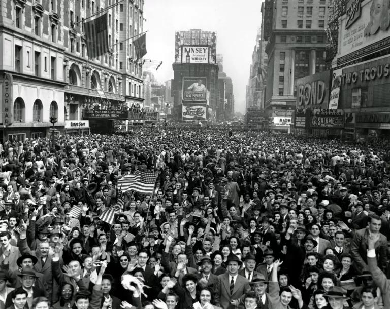 A black and white image of crowds of people packed onto a city street, some holding American flags