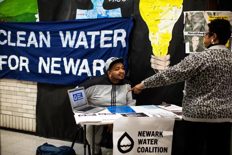 A man sits behind a table with a poster that reads "Newark Water Coalition" and a banner behind him that reads "Clean Water for Newark"