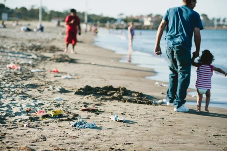 A man holds a small child's hand while walking on a beach with trash on the sand