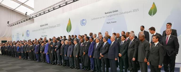 A large number of people dressed in business attire stand neatly in three rows in front of large signs with the United Nations logo on them.