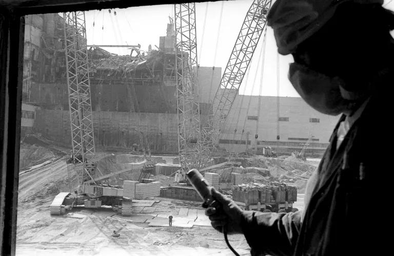 In a black and white image, a worker with protective gear reads a handheld meter, with a partially-collapsed structure in the background