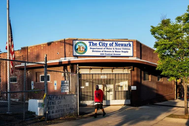 A person walks past a building with gates closed over the doors and a sign that reads in part "City of Newark Department of Water & Sewer Utilities"