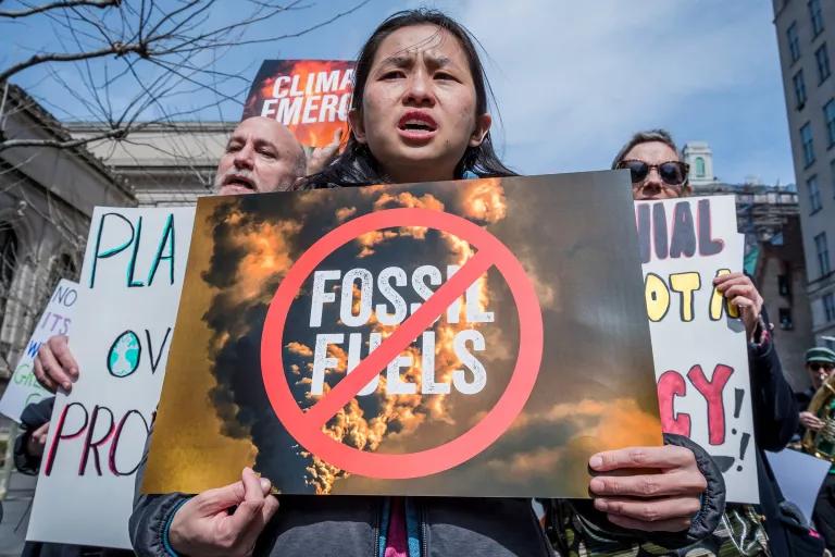 A young person carries a sign with an image of billowing smoke and the words "fossil fuels" with a red mark across them.