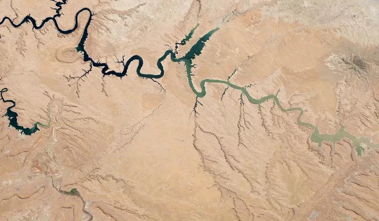 A satellite image shows a river snaking through pale brown land. The river is darker bluish-green on the left of the image and fades to a pale green on the other side of the image.