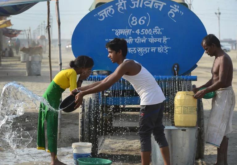 Water is dispensed from the back of a blue tanker truck. A woman splashes water on her face while one man collects water in a jug and another rinses a pot.