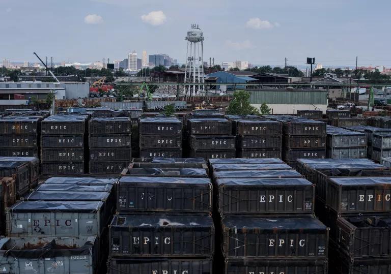 Shipping containers are stacked in long rows with a city skyline and water tower in the background