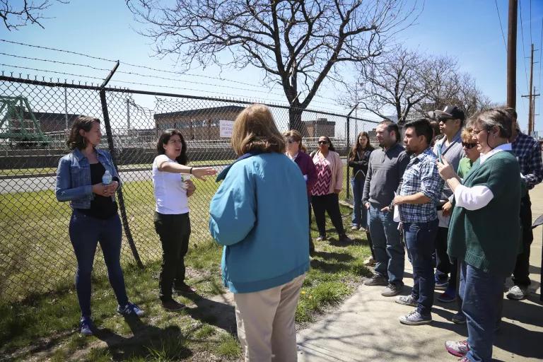 A woman stands in front of a chainlink fence and talks to a group of people