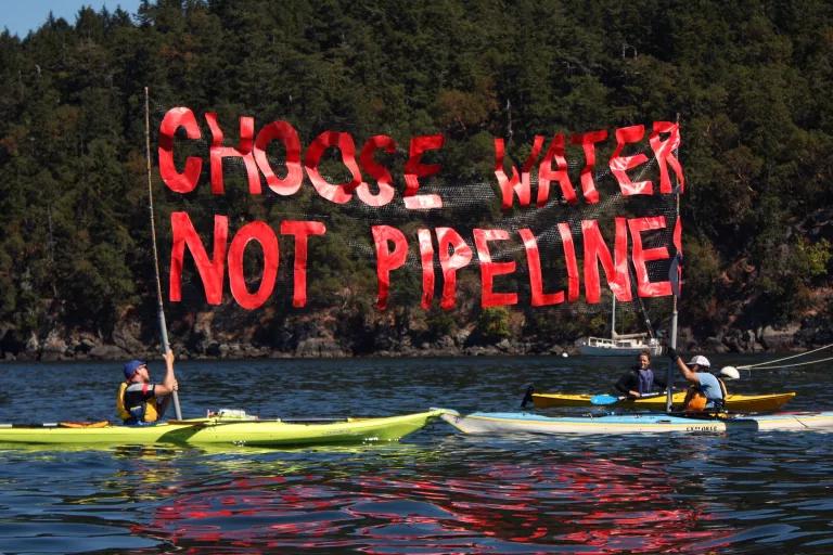 Two people in separate kayaks each hold one end of a large sign that reads "Choose Water, Not Pipeline"