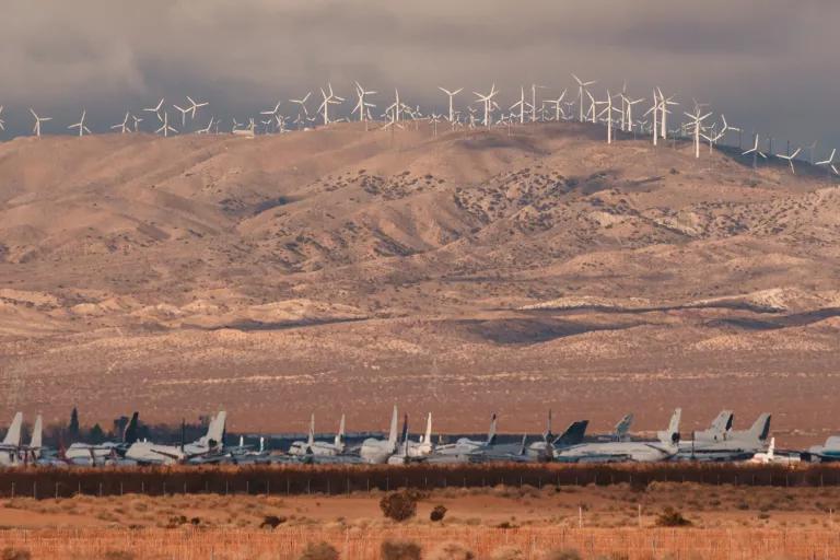 Dozens of wind turbines stand on top of a mountain with rows of military jets in the foreground
