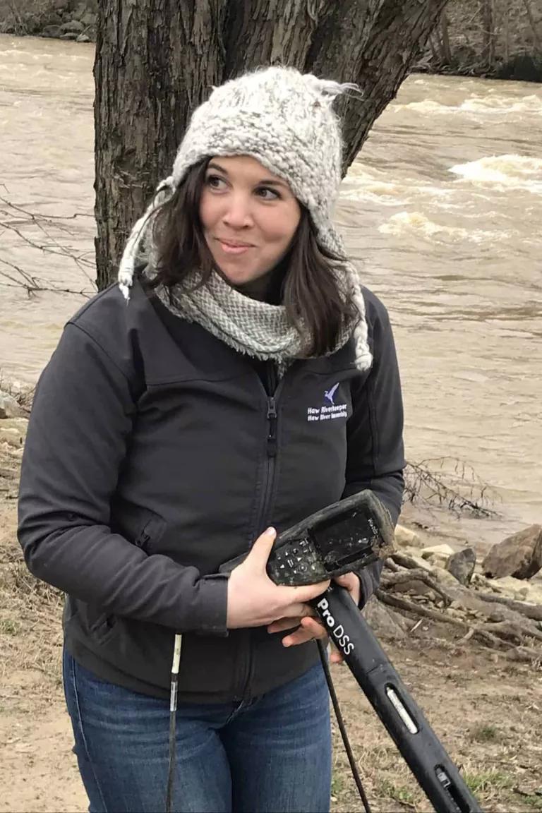 A woman standing near a riverbank carries equipment