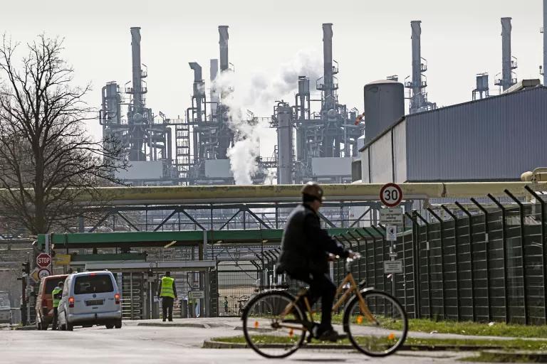 A person on a bicycle on a city street with refinery stacks rising into the sky in the background