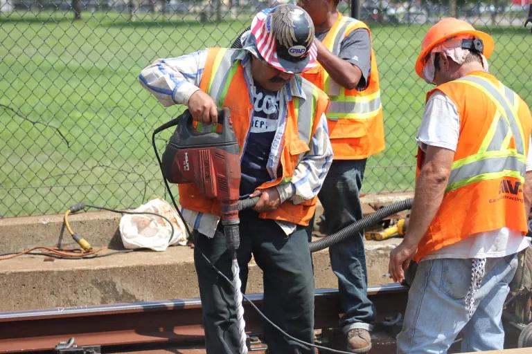 Three men in bright orange safety vests and hard hats repair a track in the sun