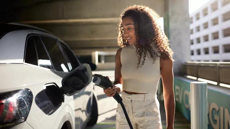 A woman holding a charger beside an electric car at a charging station in a public parking garage in Los Angeles, California.