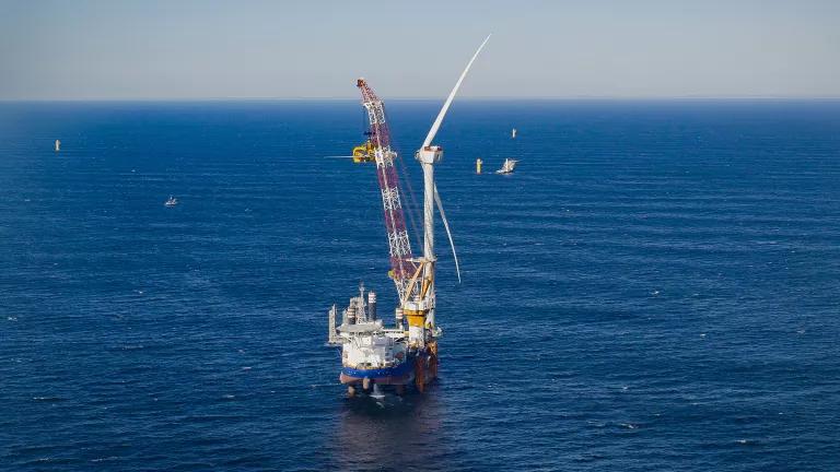A construction crane lifts a large turbine blade to be attached to an offshore wind turbine