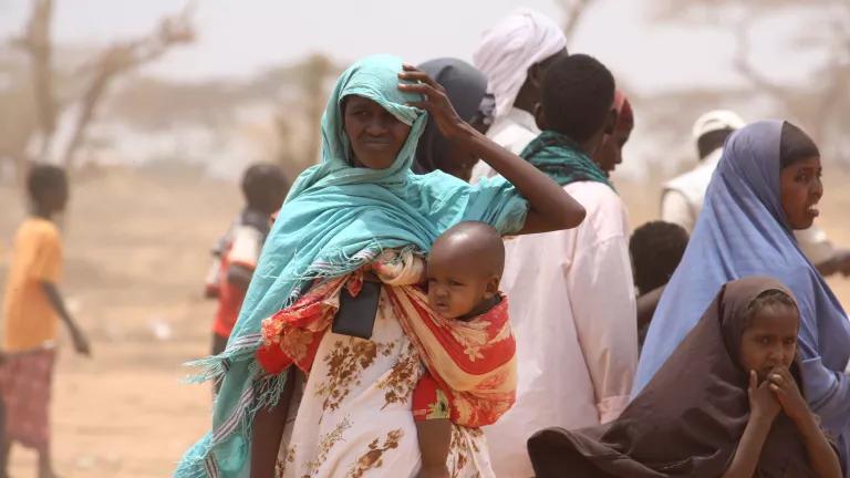 A woman carrying an infant and squinting in the sun stands with other women and children
