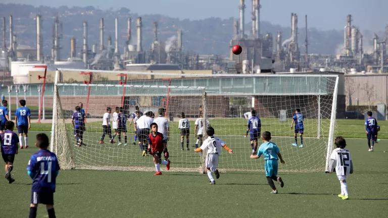 Players on a soccer field with industrial stacks rising in the background