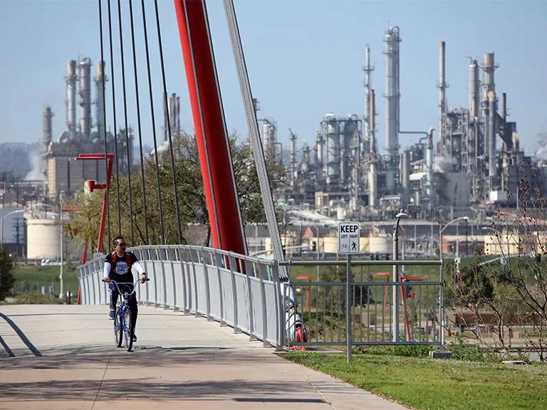 A person rides a bicycle on a paved path on a bridge with an oil refinery in the background
