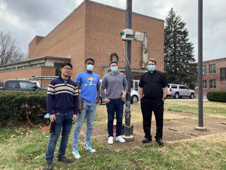 Group photo of David Yeom, Tyler Cargill, Li Zhiyao, and Reverend Nick Winker standing next to an air pollution monitor mounted on a pole in front of the St. Ann Catholic Church in St. Louis, Missouri