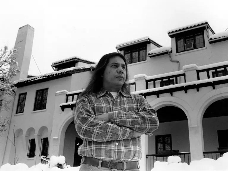 Black and white photo of John Echohawk standing with arms crossed in front of the Native American Rights Fund in the snow