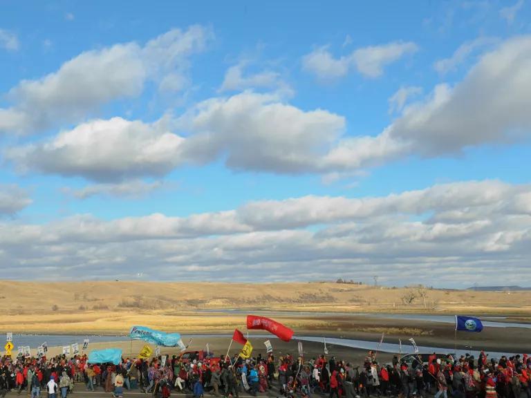 Hundreds of people march along a road and waterway, with many holding messages against DAPL.