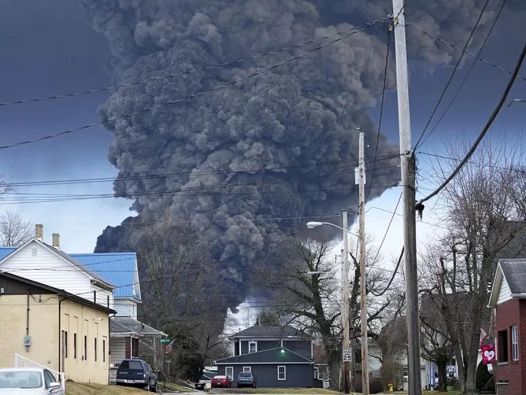 A plume of thick black smoke rises over homes on a residential street.