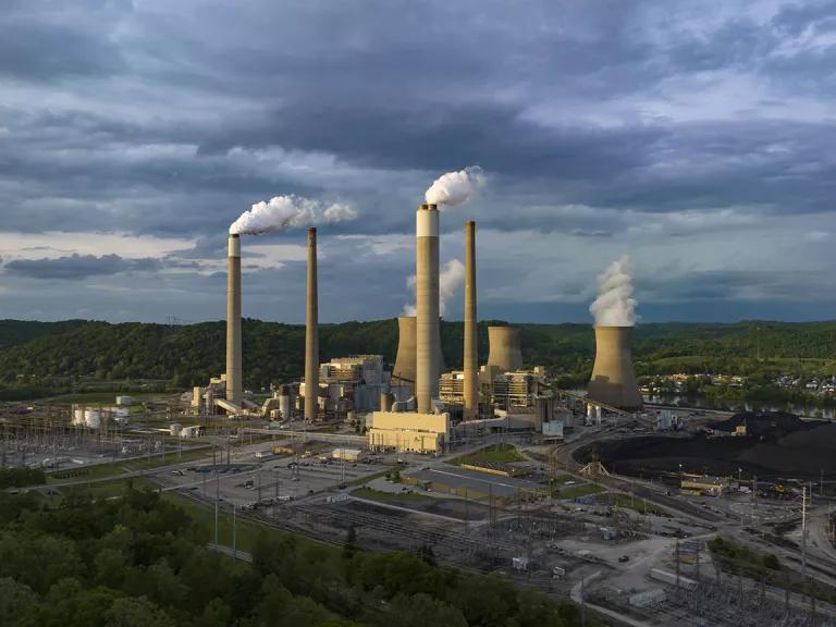 A power plant with tall stacks with billowing smoke sits in a green valley surrounded by low mountains