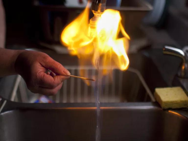 A person holds a lit match under running water, with flames rising up above the faucet