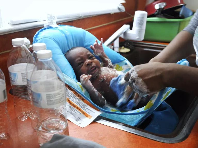 A woman washes a baby in an infant bath seat in a kitchen sink, with empty water bottles in the foreground.