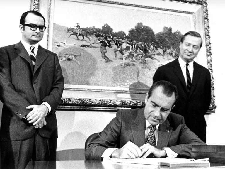 A black and white image shows President Richard Nixon sitting at a desk while two men stand on either side