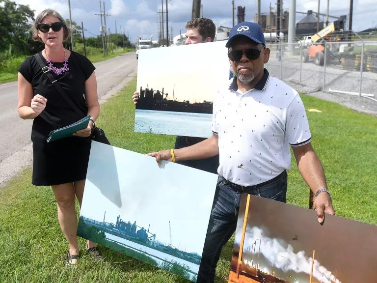 Three people stand in front of a large industrial facility and hold posters with images on them