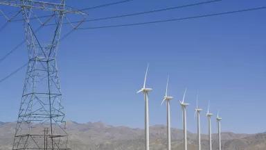 Six wind turbines stand in a row next to an electrical transmission tower, with low mountains in the background