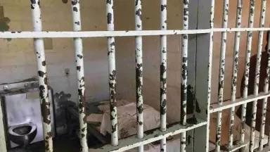 An image of a prison cell. The bars are severely rusted. There is a combination sink-toilet fixture visible on the left side of the image and a cot on the right side, along with a small electric fan.