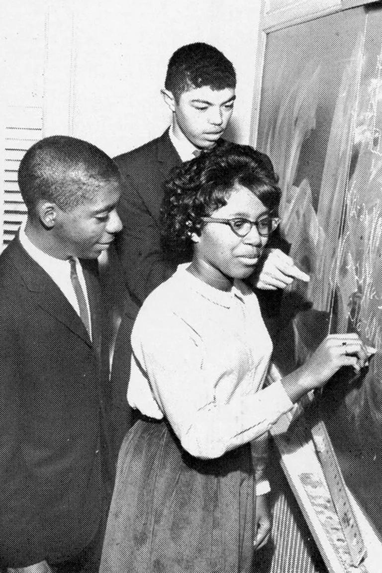 A black-and-white image of a woman writing on a chalkboard with two men looking on