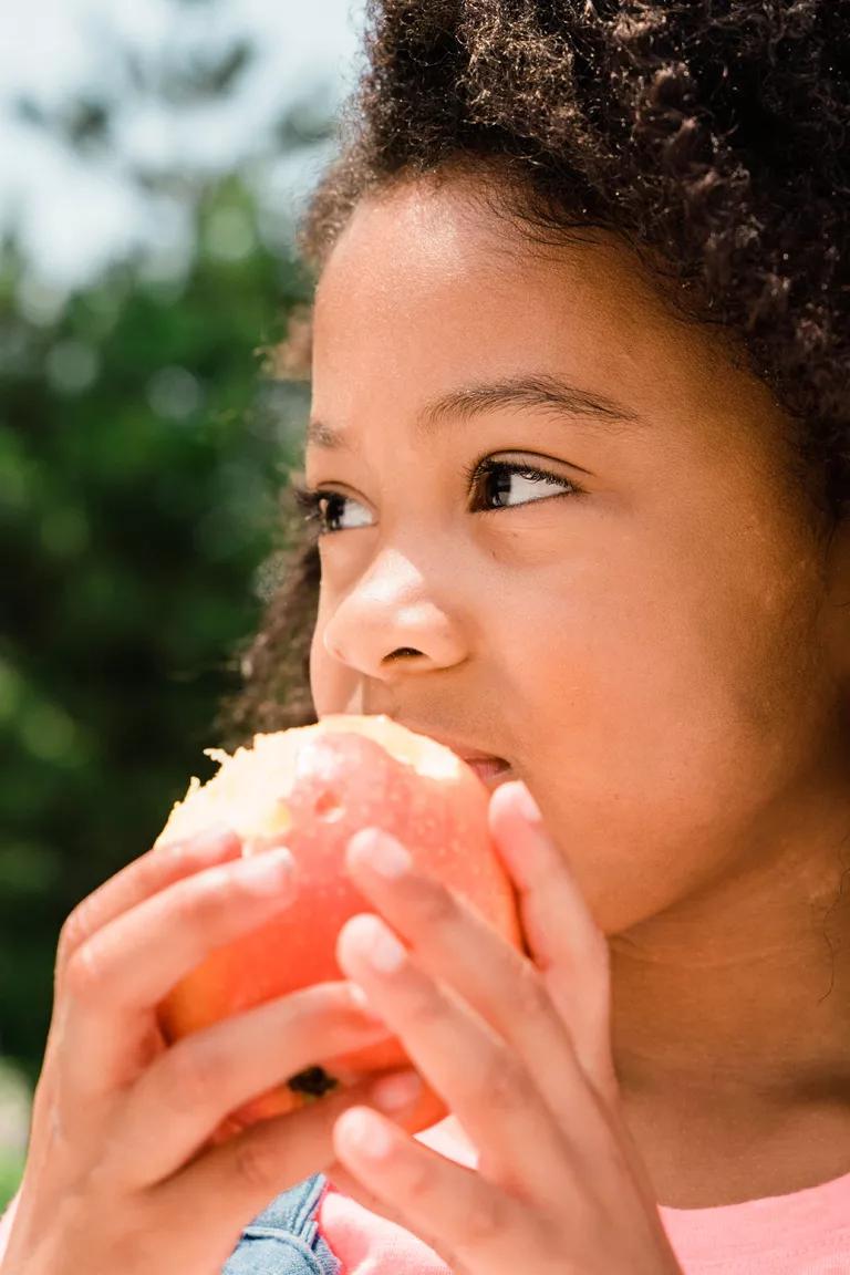 A close-up of a child's face, who's eating an apple with both hands, with green trees in the background