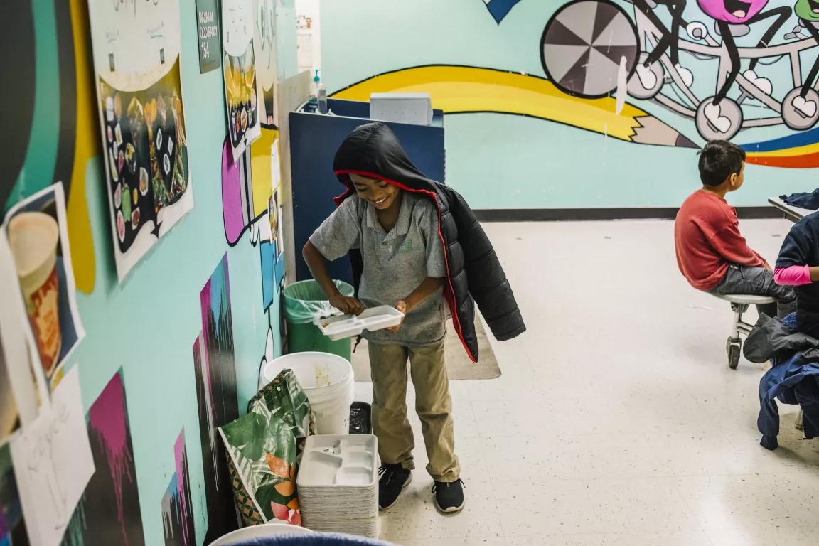 A child in a school cafeteria empties a lunch try into bins along the wall