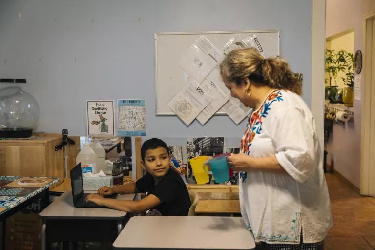 A woman hands a cup of water to a child sitting at a desk working on a laptop