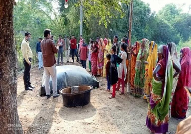 A group of primarily female villagers in Aravalli District, Gujarat stand in front of a biogas plant installation. A man faces them, gesturing about the benefits of biogas.