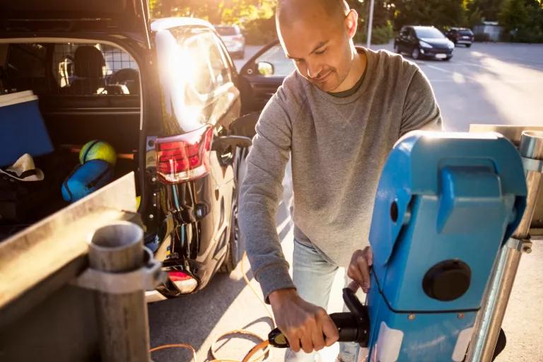 A smiling person in a grey shirt and blue jeans is plugging in their car to charge in an outdoor parking lot. The car's trunk is open and has sports equipment inside.
