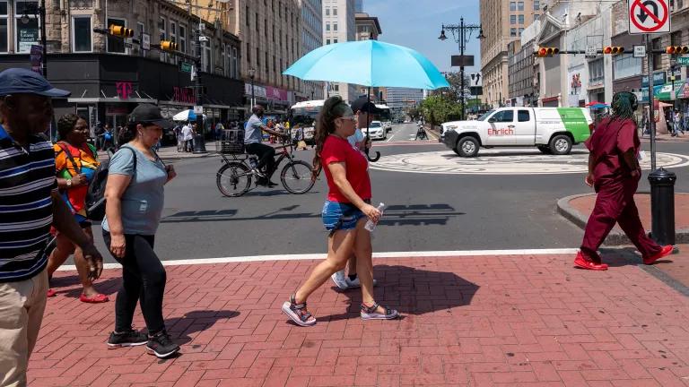 Pedestrians walking on a sunny city street, one woman holding an umbrella and carrying a water bottle.