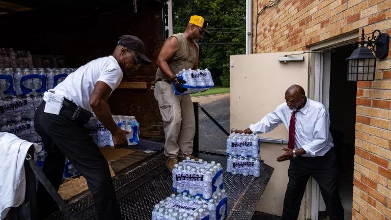 Three men unload cases of water from the back of a delivery truck and into a brick building