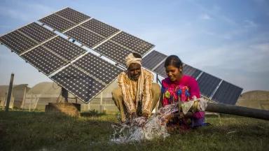 Two women kneel down at a water pump with a solar panel in the background