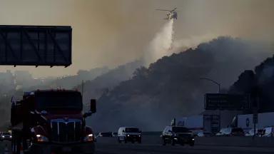 Trucks and cars driving on a freeway as a helicopter drops water on fast-moving brush fire.
