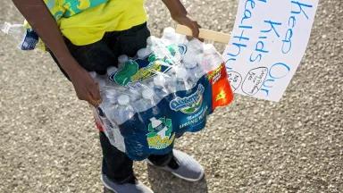 A person wearing a yellow t-shirt holds a case of bottled water and a poster that reads "Keep our kids healthy"
