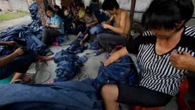 Workers in a shed sit on the floor and low stools while hand-sewing denim jeans