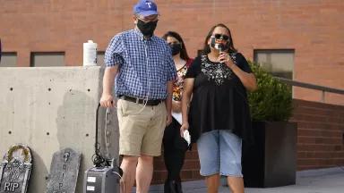 A man and woman wearing facemasks stand with a portable microphone and speaker in front of a brick building 