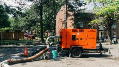 A large orange pump with large hoses attached sits on a trailer parked in a concrete lot with a brick apartment building in the background